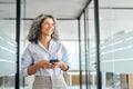 Happy mature business woman using mobile phone standing in office looking away. Royalty Free Stock Photo