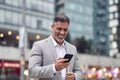 Happy middle aged businessman wearing earbud using phone walking on city street.