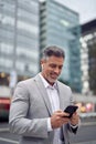 Happy middle aged businessman wearing earbud using phone walking on city street.