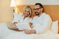 happy middle aged couple reading book and using digital tablet while lying together in bed Royalty Free Stock Photo