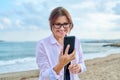 Happy middle aged woman on sea vacation enjoying in headphones with smartphone