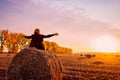 Happy middle-aged woman sitting on haystack in autumn field and feeling free with arms opened