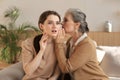 Happy middle mother whispering secret to her smiling daughter at home, gossiping, sharing secret