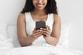 Happy millennial african american lady relaxing in comfortable bed, holding smartphone in hands, crop