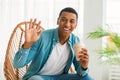 Happy millennial african american man with cup of coffee takeaway and waving hand, sits on chair, enjoys comfort