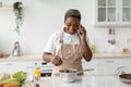 Happy millennial african american woman in apron prepare food and making phone call in modern scandinavian kitchen Royalty Free Stock Photo