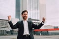 Happy millennial bearded businessman in suit rejoices in successful deal or win, rises hands up near modern office