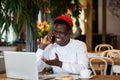Happy millennial Black man laughing, speaking with friend on mobile phone, remote work in cafe Royalty Free Stock Photo