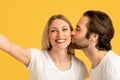 Happy millennial caucasian husband kissing wife on cheek in white t-shirt, lady make selfie