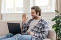 Happy millennial caucasian man with stubble sits on sofa waving hand and looking at laptop in living room