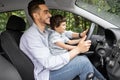 Happy millennial dad teaching little boy with steering wheel to drive car in summer, outdoor, profile Royalty Free Stock Photo