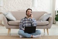 Happy millennial european guy with stubble in shirt calls by phone, works on laptop, sits on floor in living room