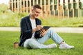 Happy millennial european man enjoy lifestyle, work on tablet with coffee cup takeaway, sit on grass in city Royalty Free Stock Photo