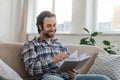 Happy millennial handsome caucasian guy with stubble sits on sofa reads book in living room interior