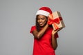 Happy african woman in Santa hat holding Christmas present, going to open gift, grey studio background