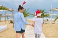 Happy mom and daughter child in Santa hat walking along beach, back view Royalty Free Stock Photo