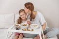 Happy mom and daughter have healthy breakfast on bed in a light bedroom on a sunny morning. Mom kisses daughter. Healthy food and