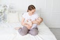 Happy mom holds baby in her arms with a rodent in her mouth on a white bed with cotton bedding at home