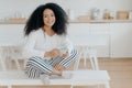 Happy morning concept. Photo of cheerful curly African American woman sits in lotus pose at white bench, sips tasty aromatic drink Royalty Free Stock Photo