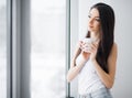 Happy morning. Portrait of beautiful young woman standing at window, drinking coffee and looking outside enjoys of rest Royalty Free Stock Photo