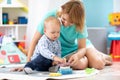 Happy mother and adorable baby boy playing on floor mat in sunny nursery room. Cute child with carer in creche