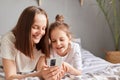 Happy mother and cute daughter using mobile phone, resting on bed at home, little girl with mommy looking at cell phone display Royalty Free Stock Photo