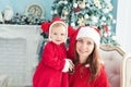 Happy mother and daughter in Christmas hats and in red dress sitting in armchair near fireplace and Christmas tree with gifts Royalty Free Stock Photo