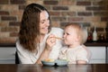 Happy mother feeding her kid puree mixture from a spoon in kitchen. Healthy eating, baby weaning. Complementary feeding