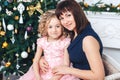 Happy mother with her daughter sitting near a Christmas tree looking into the camera and smiling