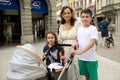 Happy mother with kids walk with baby pram, stroll the Italian medieval city during family holidays, smiling at camera