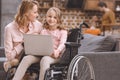 happy mother looking at cute little daughter sitting in wheelchair and using laptop