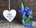Happy Mother`s Day greeting card with decorative white heart and blue spring flowers in a glass vase on old wooden background. Royalty Free Stock Photo