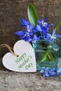 Happy Mother`s Day greeting card with decorative white heart and blue spring flowers in a glass vase on old wooden background. Bea Royalty Free Stock Photo