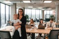 Happy mother is standing and holding her little son. Infant baby is in the office where group of people are working together Royalty Free Stock Photo