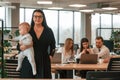 Happy mother is standing and holding her little son. Infant baby is in the office where group of people are working together Royalty Free Stock Photo