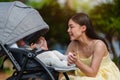 happy mother talking and playing with her infant baby in the stroller while resting in park