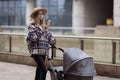 Happy Mother walking With Stroller In Park and using mobile phone. Joy of motherhood. Stylish young caucasian woman Royalty Free Stock Photo