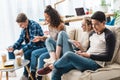 happy multicultural teens sitting on sofa with digital