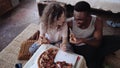 Happy multiethnic couple sitting on the flow, hugging and eating fast food. Woman feed the hungry man a slice of pizza. Royalty Free Stock Photo