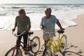 Happy multiracial senior couple talking while wheeling bicycles on shore at beach during sunny day