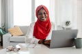 Happy muslim african american woman sitting at workplace and smiling at camera while working on laptop at home office Royalty Free Stock Photo