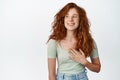Happy natural girl with red hair and freckles, holding hand on heart, smiling and looking asie with cheerful face