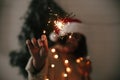 Happy New Year eve party atmosphere. Sparkler burning in hand of stylish girl in santa hat on background of modern christmas tree Royalty Free Stock Photo