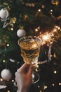 Happy New Year! Hand holding champagne glass with burning sparkler on background of christmas tree lights. Firework bengal light Royalty Free Stock Photo