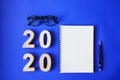 Happy new year 2020 year, with plywood number 2020, notebook, black vintage glasses and a pencil on blue paper background Royalty Free Stock Photo