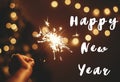 Happy New Year text sign on glowing sparkler in hand on background of golden christmas tree lights in dark festive room. Fireworks Royalty Free Stock Photo