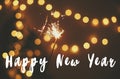 Happy New Year text sign on glowing sparkler in hand on background of golden christmas tree lights in dark festive room. Fireworks Royalty Free Stock Photo