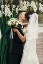 Happy newlywed couple hugging under white flowers aisle arch, handsome groom kissing beautiful brunette bride at wedding ceremony Royalty Free Stock Photo