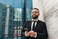 Happy office worker using smartphone and listening music in wireless earphones, standing near modern office building Royalty Free Stock Photo