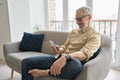 Happy older middle aged man using phone relaxing sitting on couch at home.